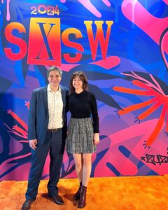 Nicole Mirra and Antero Garcia standing together in front of a blue and pink abstract backdrop that reads: SXSW 2024. The carpet is a bright orange color.